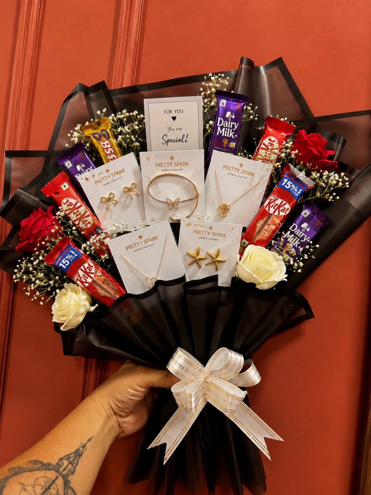 Gift bouquet with gold jewelry, chocolate bars, and roses wrapped in black paper.