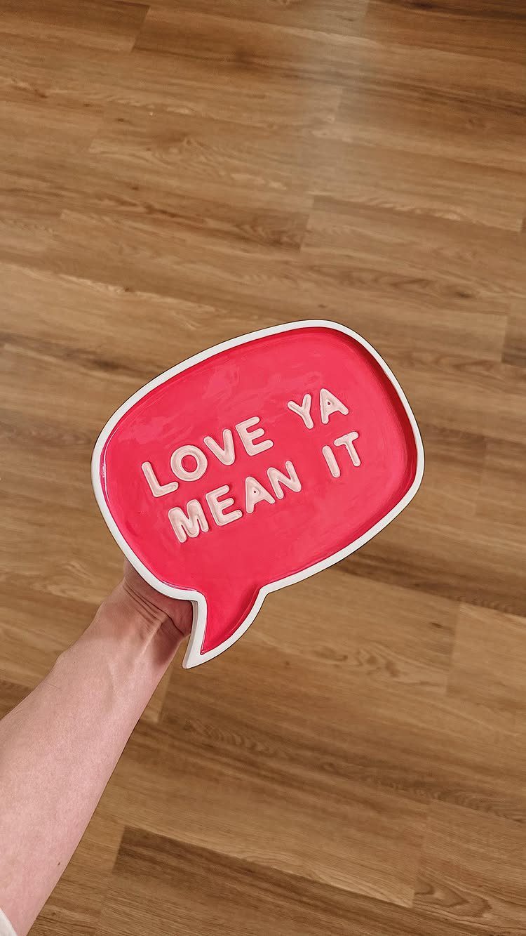 Red speech-bubble shaped ceramic dish with white text held over wooden floor.