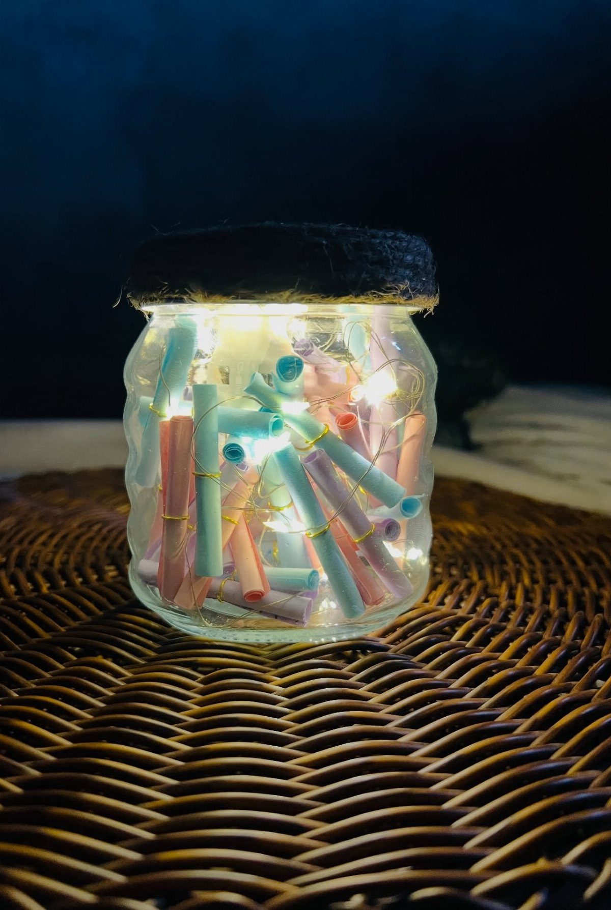 Glass jar filled with pastel paper scrolls and glowing fairy lights on wicker.