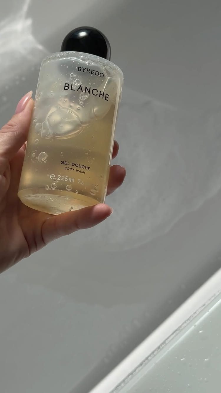 Byredo Blanche body wash bottle held over a soapy white bathtub