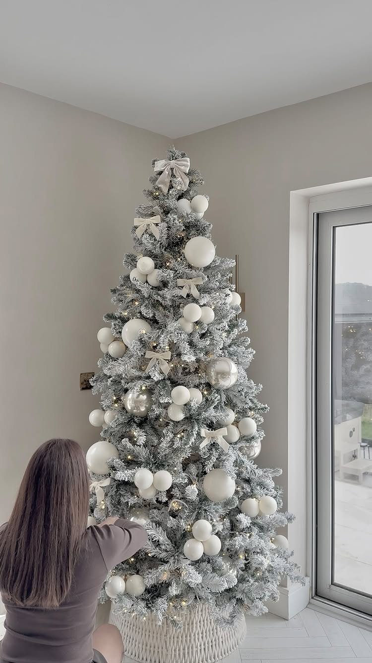 Flocked Christmas tree decorated with white ornaments and bows in a bright room.
