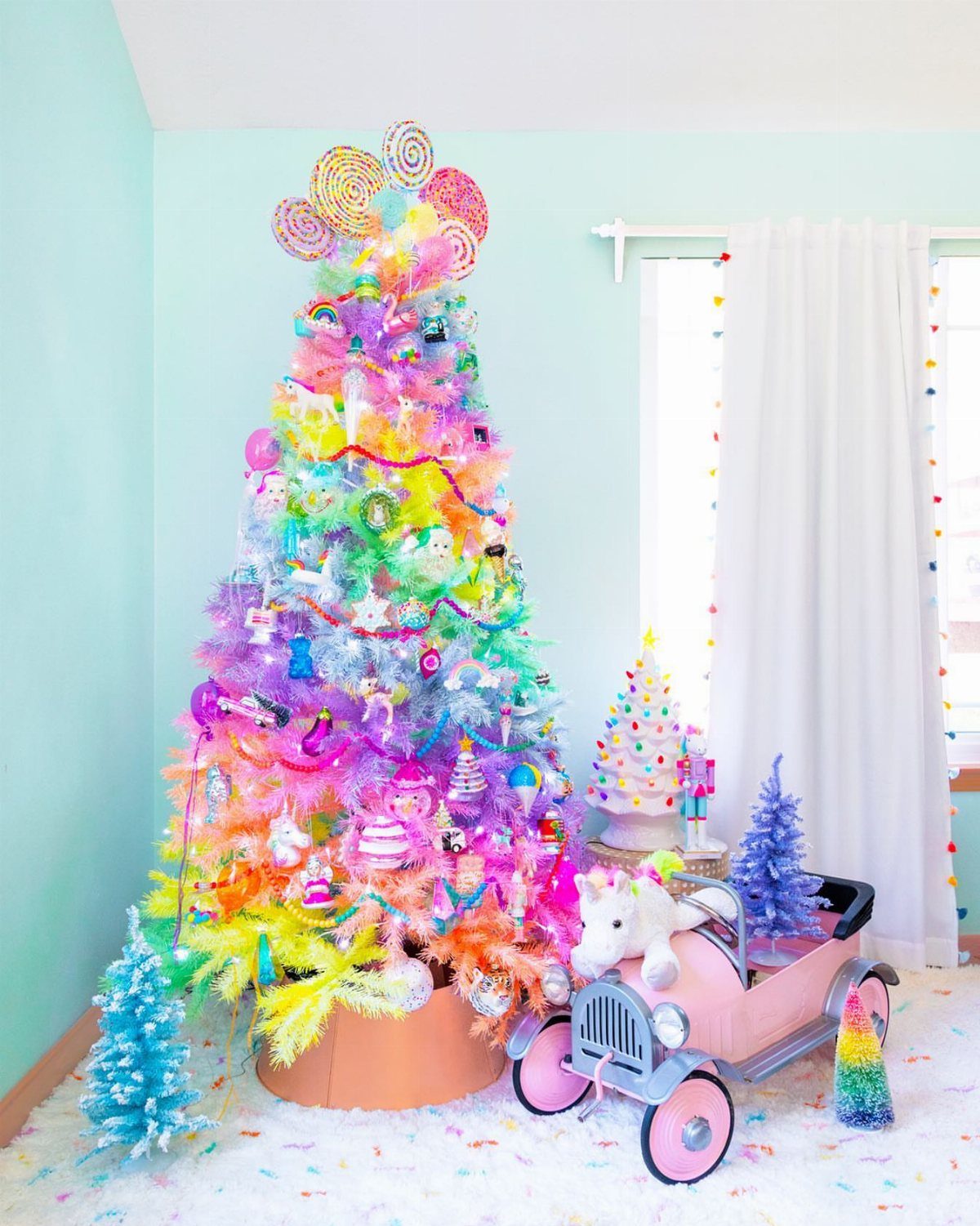 Rainbow decorated white Christmas tree with colorful ornaments and a pink toy car.