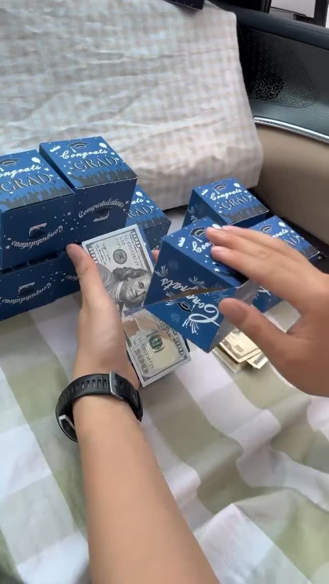Hands holding a blue graduation gift box and cash on striped fabric.