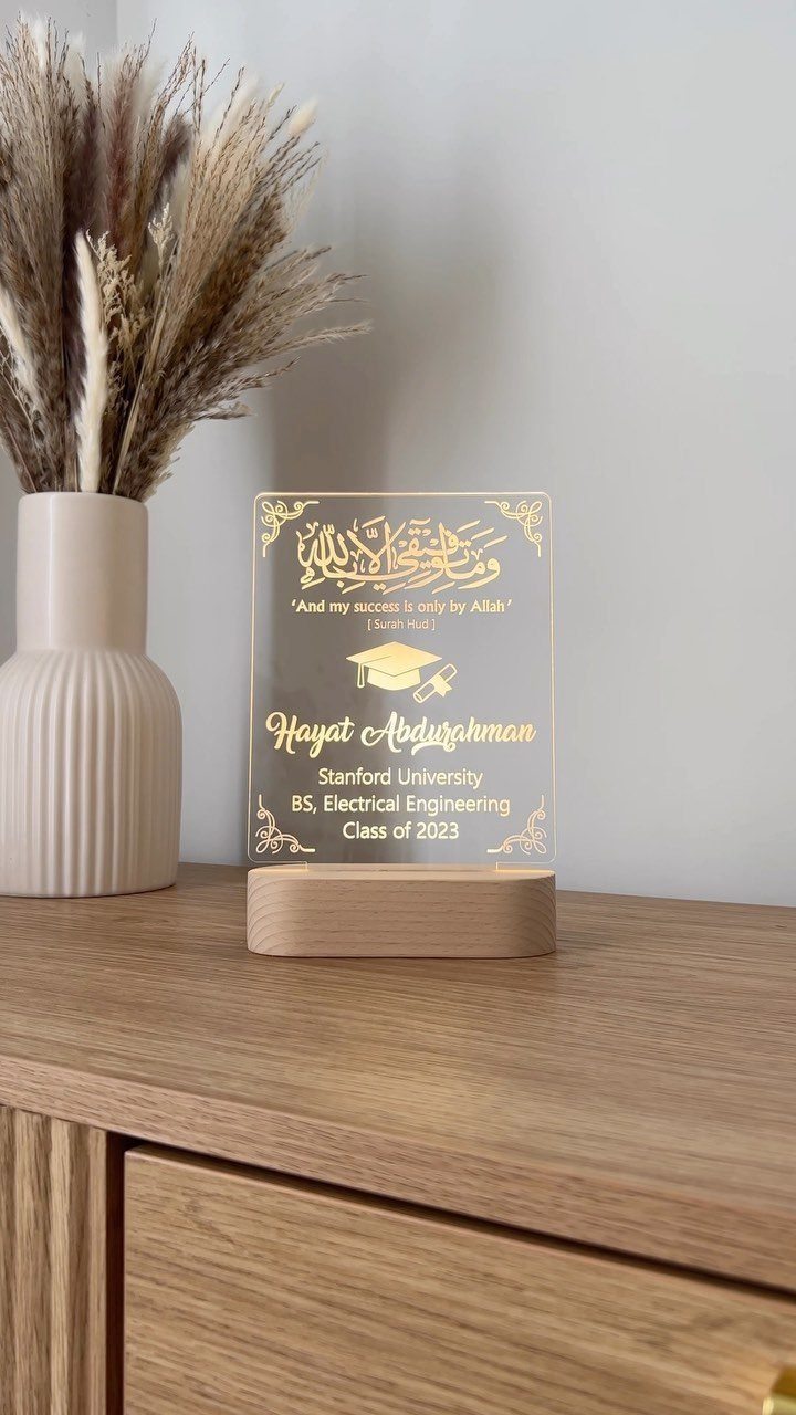 Engraved acrylic graduation plaque on a wooden base next to dried pampas grass.
