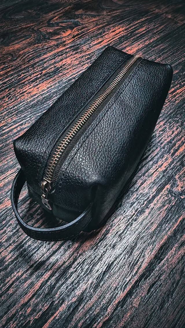 Black pebbled leather toiletry bag with gold zipper on a dark wooden surface