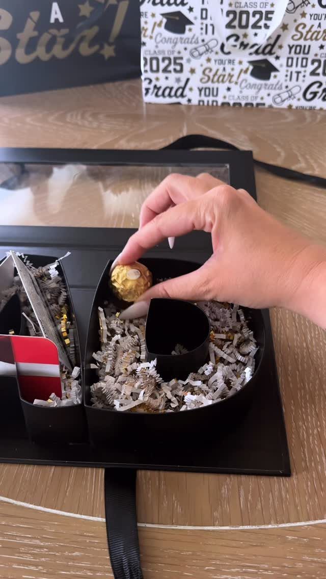 Hand placing a treat into a black letter-shaped graduation gift box with shredded paper.