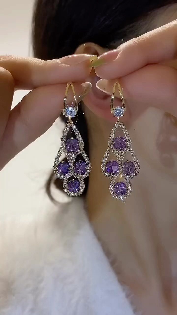 Gold drop earrings with purple gemstones and crystals held up to an ear.