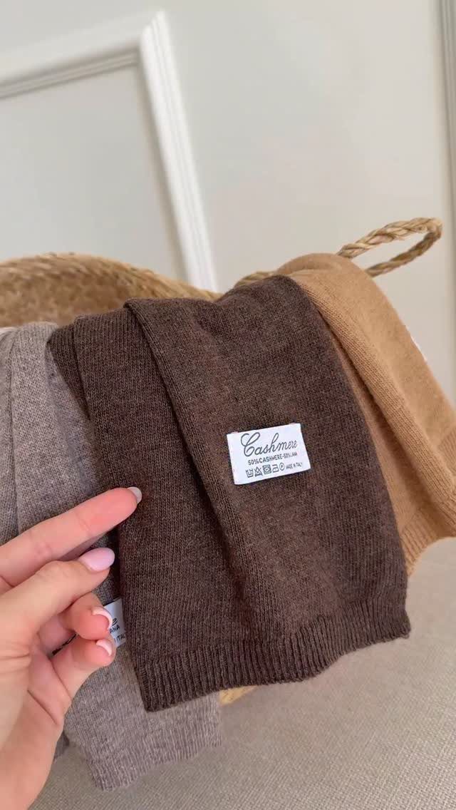 Brown cashmere sweater held up from a basket of folded neutral knitwear.
