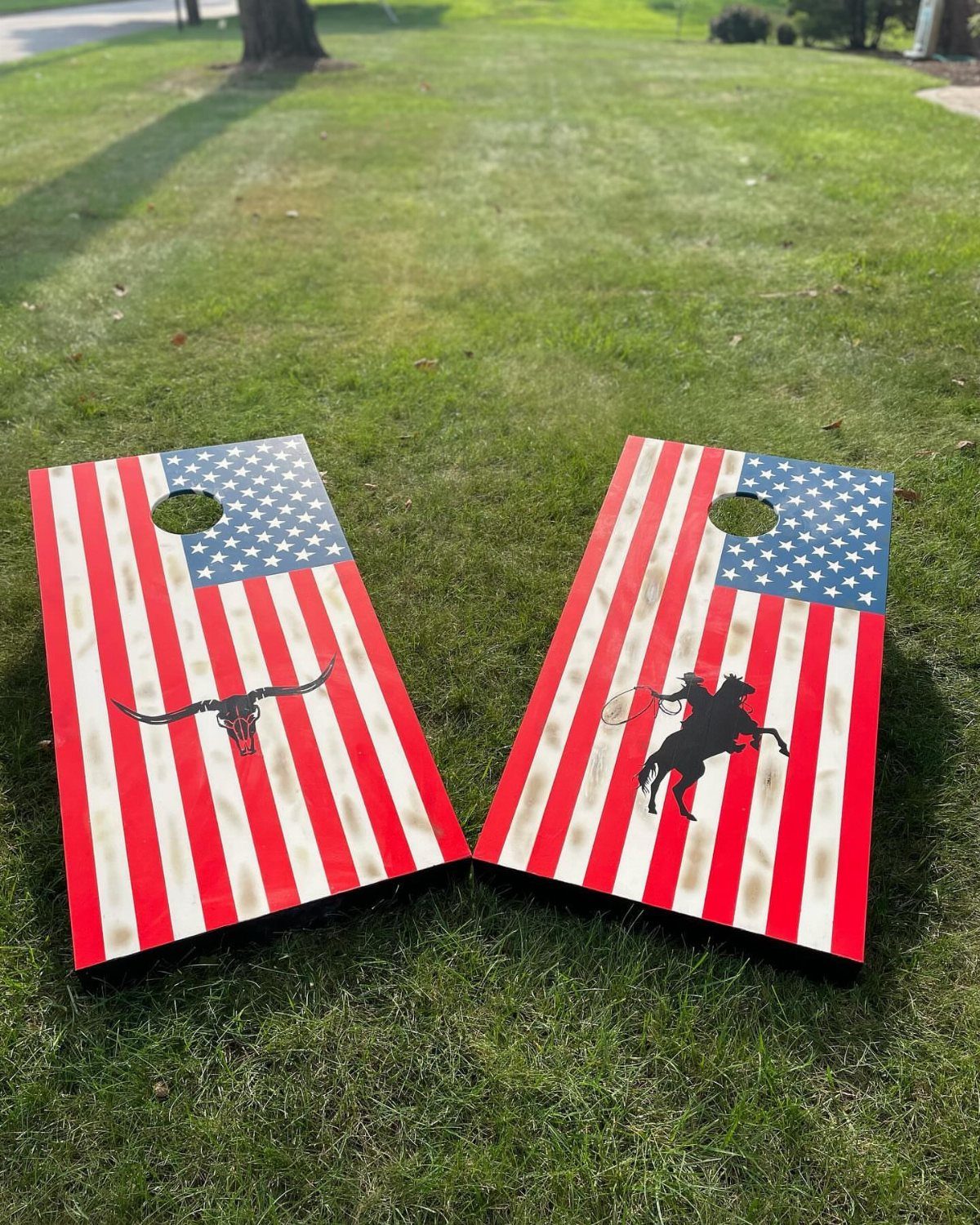 Two cornhole boards with American flag and western silhouettes on green grass.