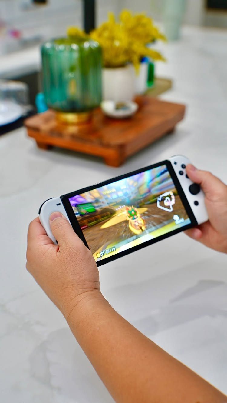 Hands holding a white handheld gaming console playing a colorful video game indoors.