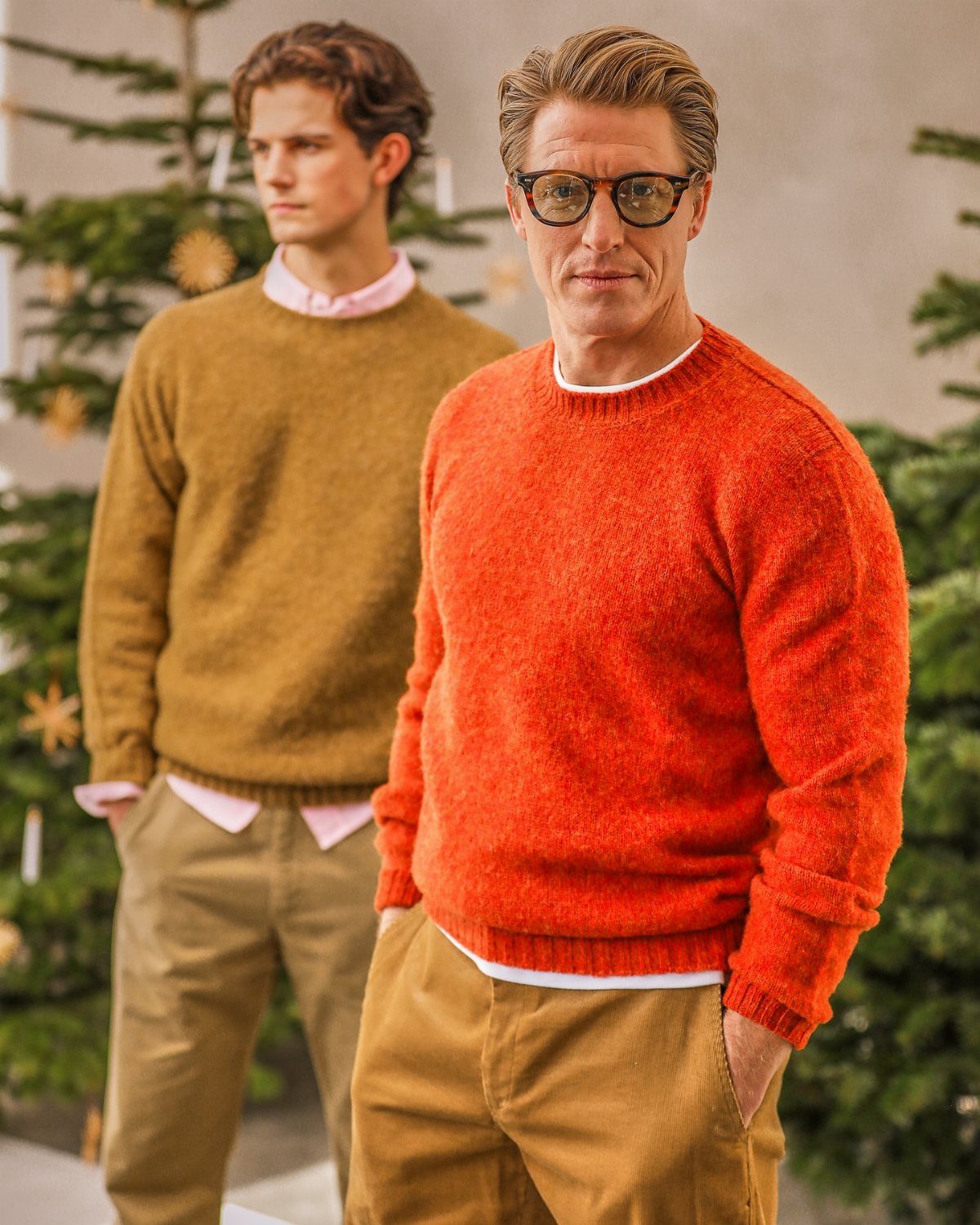 Two men wearing colorful knit sweaters standing in front of decorated Christmas trees.