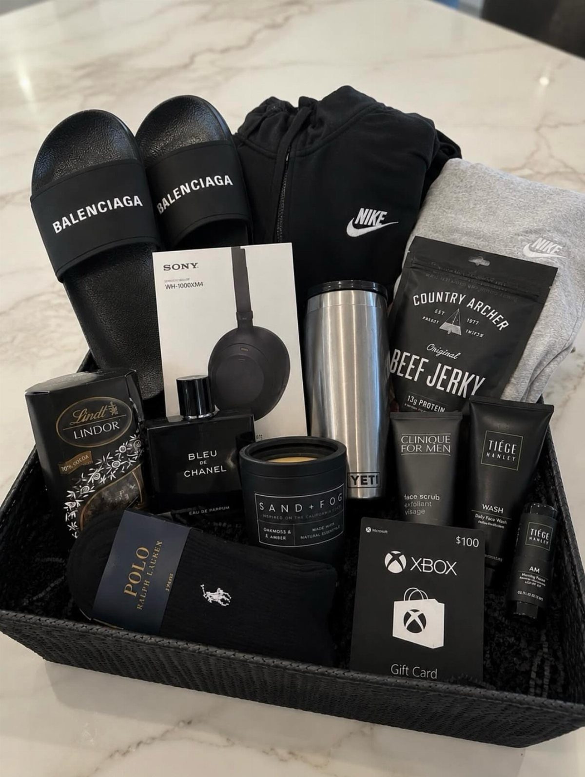 Gift basket for men containing luxury slides, electronics, clothing, snacks, and grooming products.