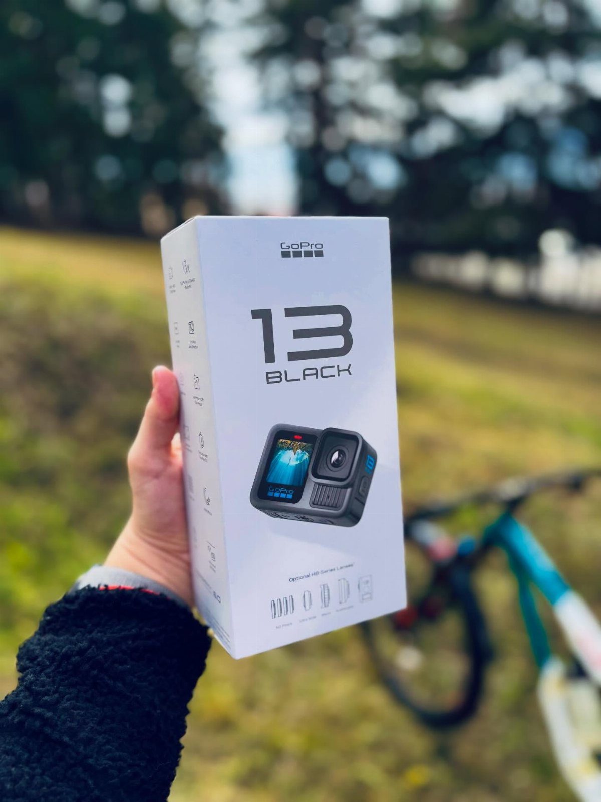 GoPro Hero 13 Black camera box held outdoors with a bicycle behind.