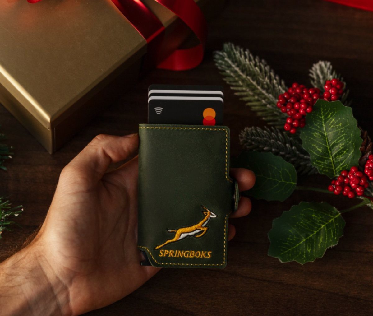 Dark green leather wallet with Springboks logo held by a hand near gift box.