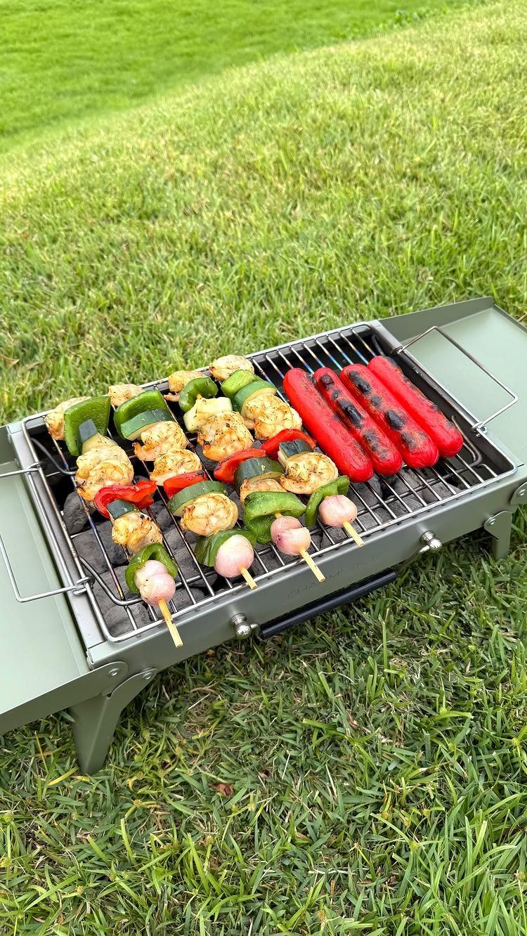 Portable olive-green grill with chicken skewers and hot dogs on green grass