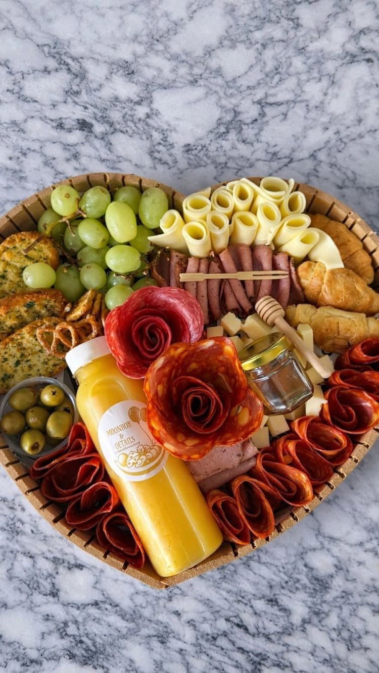 Heart-shaped gourmet gift basket with meats, cheeses, fruits, and juice on marble.
