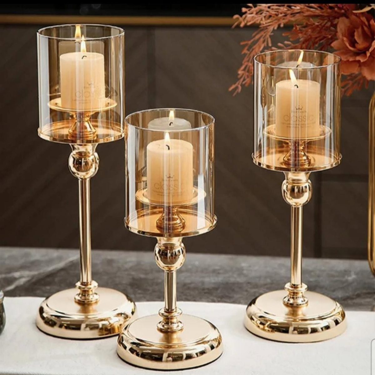 Three gold candle holders with glass cylinders and lit cream pillar candles.