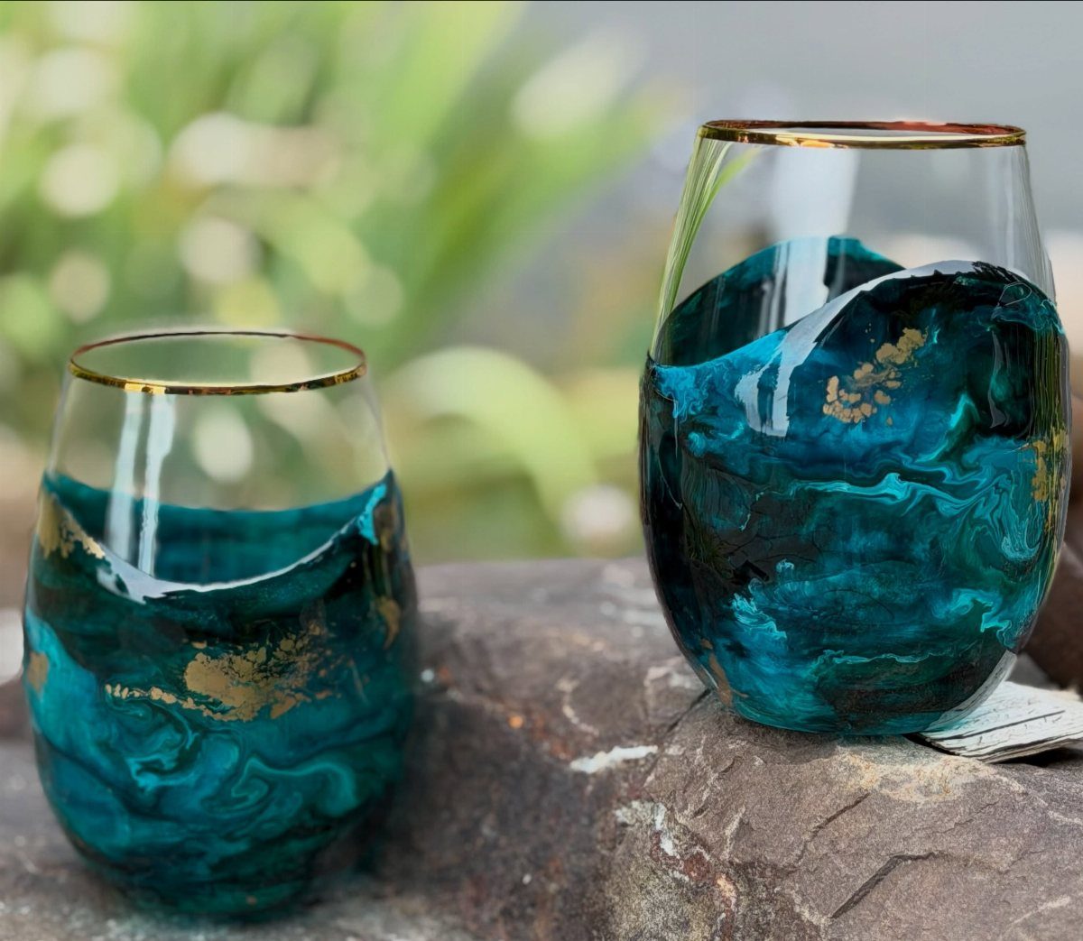 Two marbled teal and gold stemless cocktail glasses resting on a grey stone.