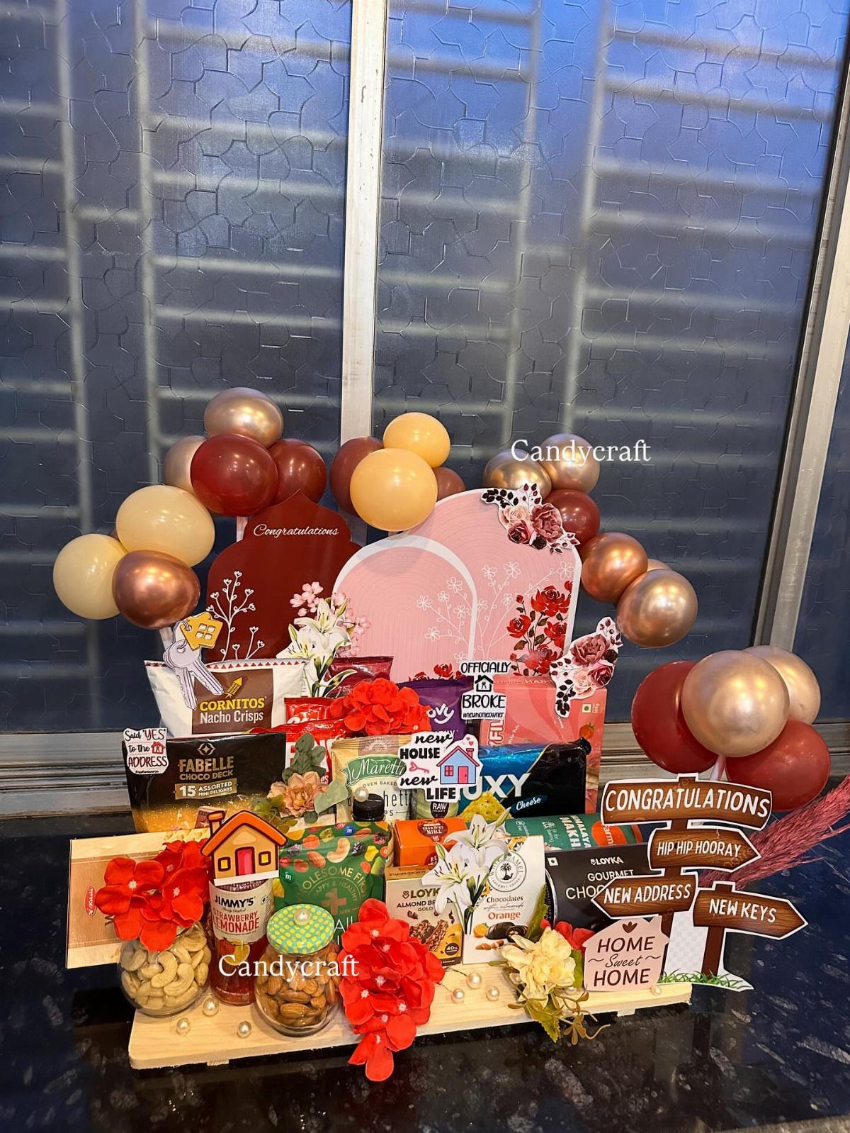 Celebratory gift basket with snacks, balloons, and housewarming signs on a wooden tray.