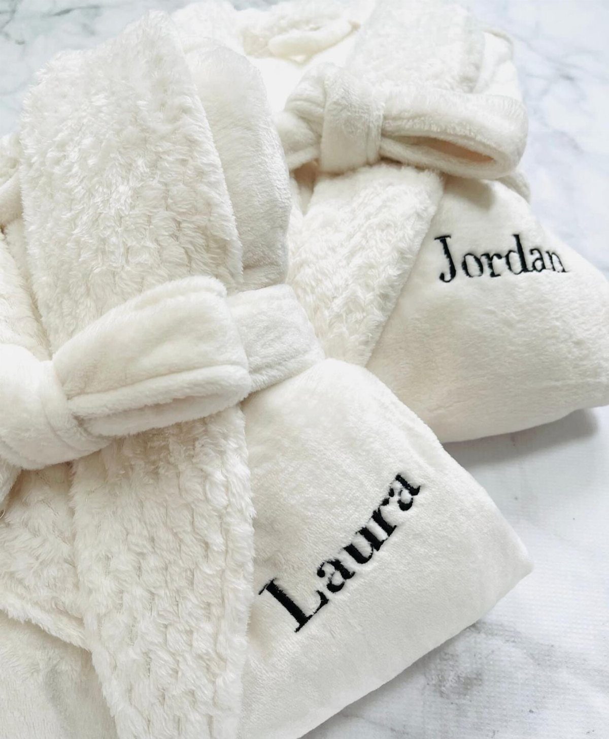 Two plush white robes with embroidered names Laura and Jordan on marble surface.