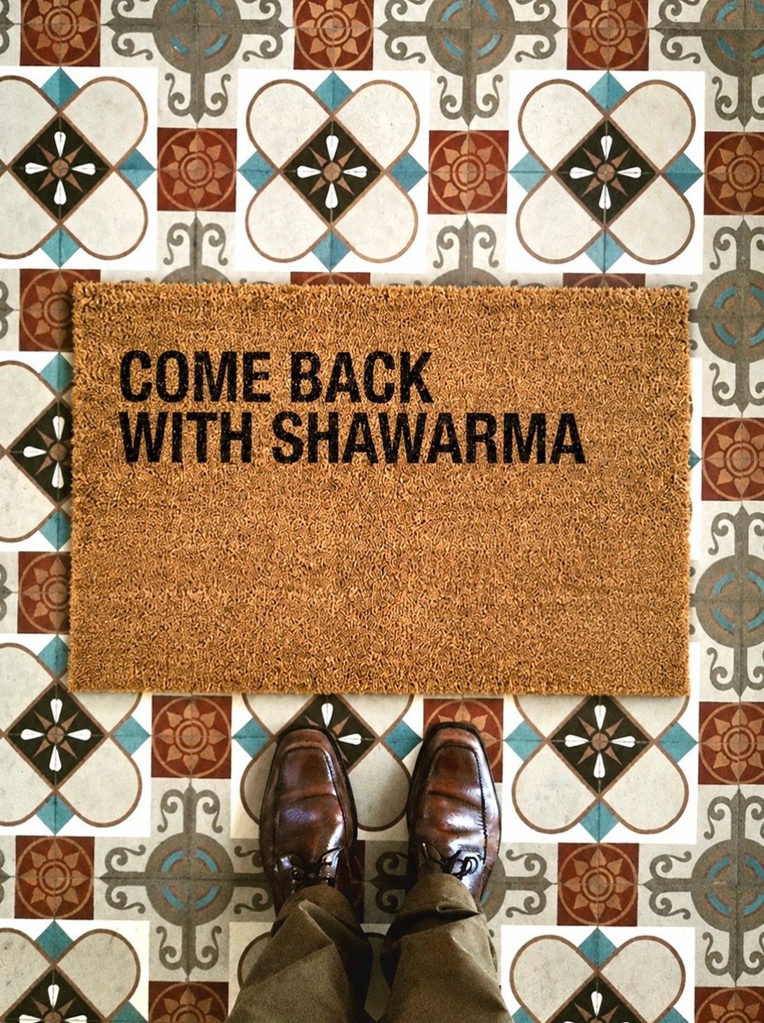 Brown coir doormat with funny text on a patterned tile floor with shoes.