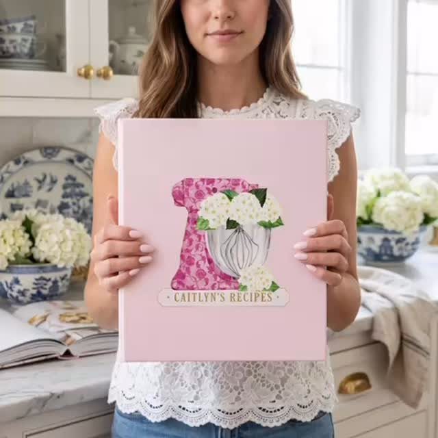 Woman holding a pink personalized recipe book in a bright, white kitchen setting.