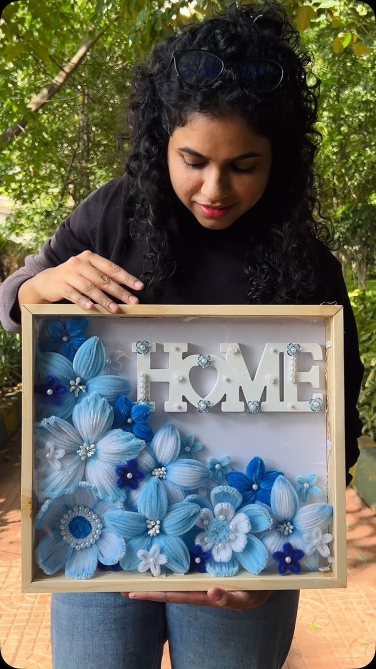 Woman holding a framed art piece with blue fabric flowers and the word HOME.