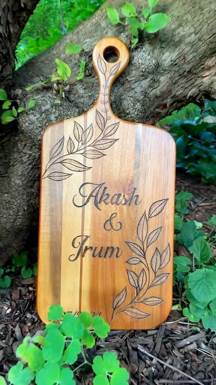 Engraved wooden cutting board with names and leaf designs leaning against a tree.