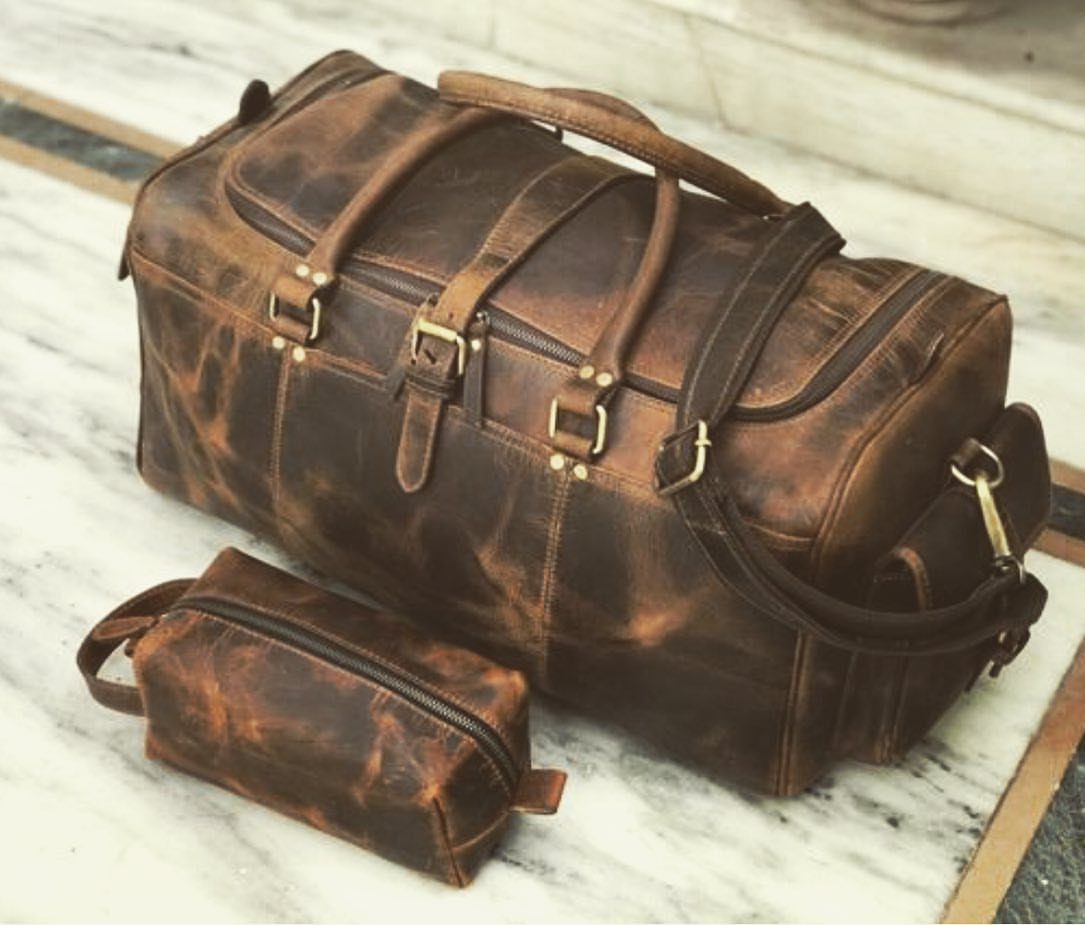 Brown leather weekender duffel bag and matching toiletry kit on white marble surface.