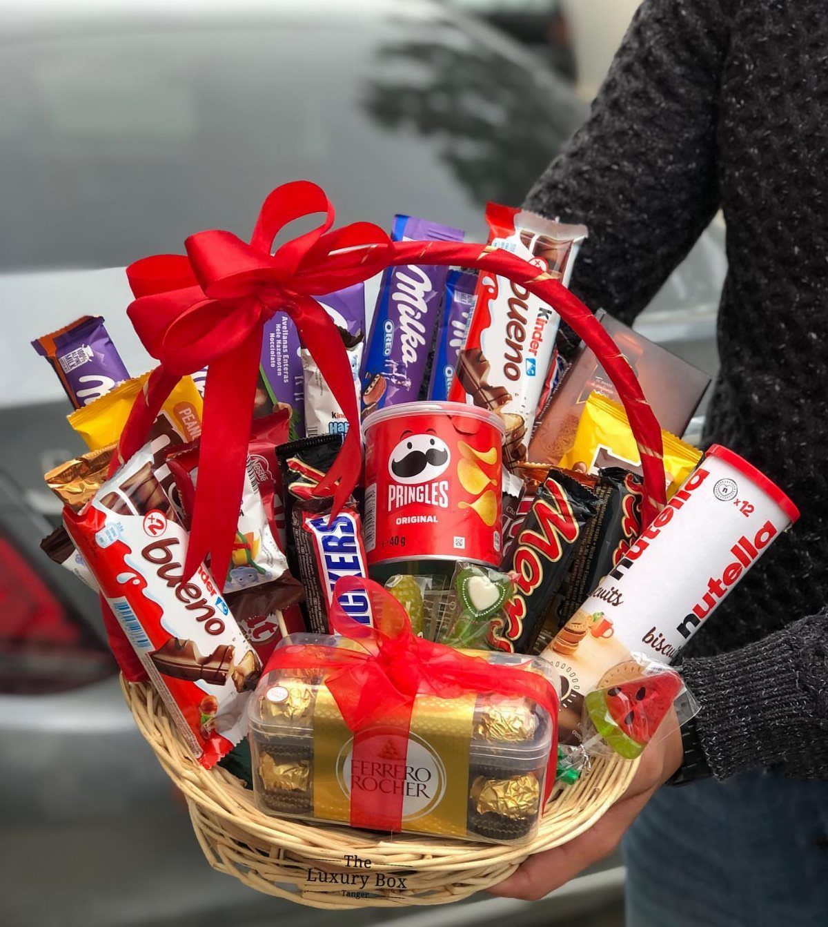 Wicker gift basket filled with assorted chocolates and snacks tied with red ribbon.