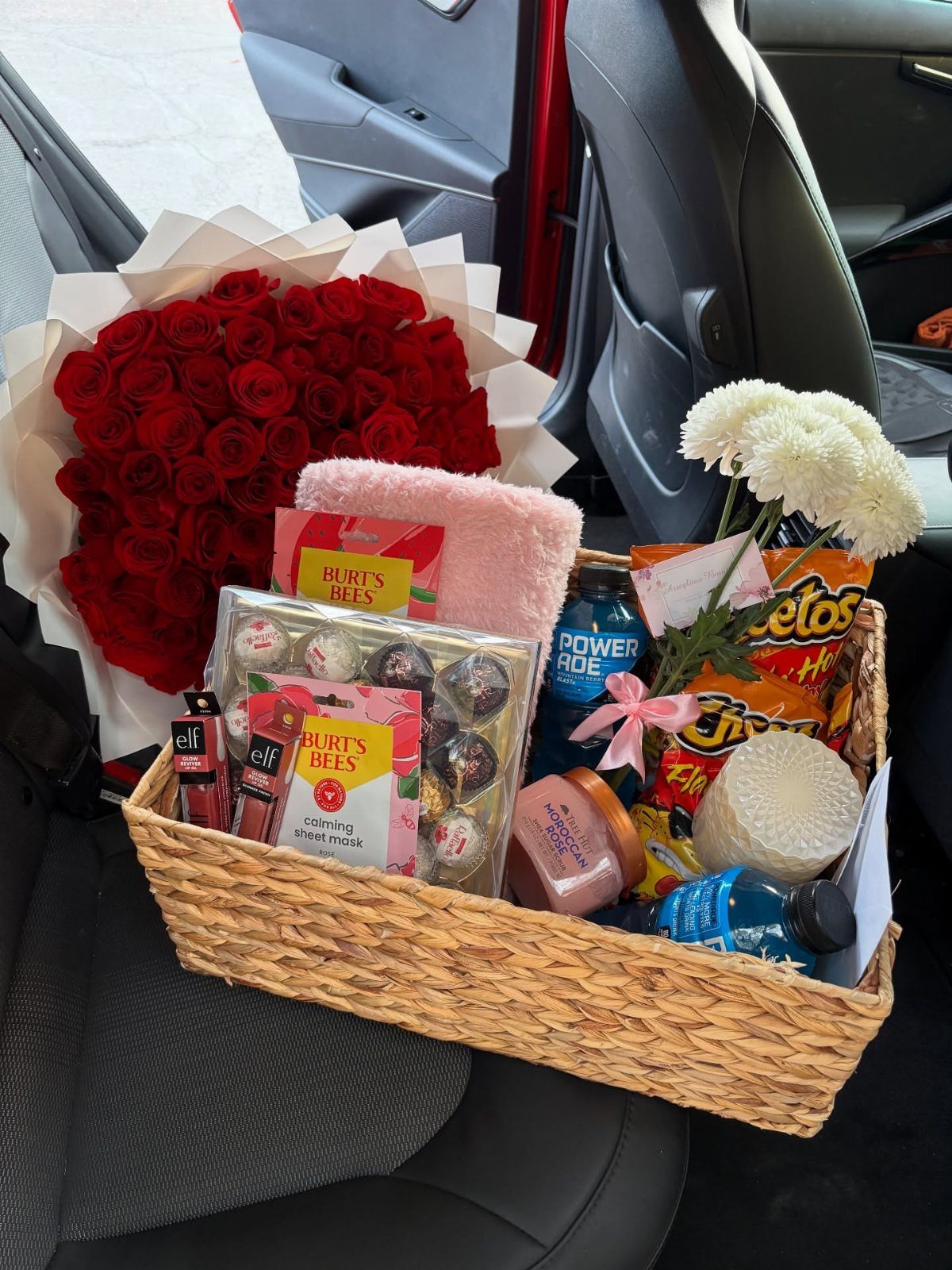 Wicker gift basket with snacks and skincare next to red roses in car.