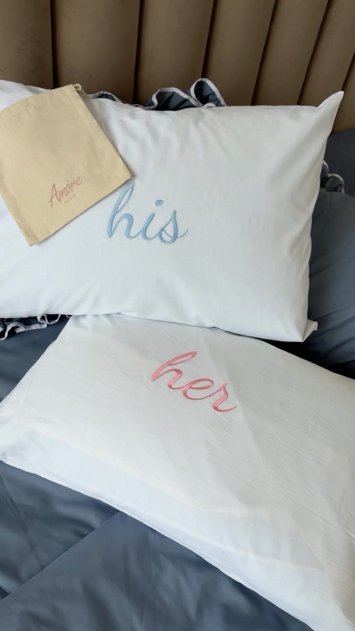 Two white pillowcases with blue 'his' and pink 'her' embroidery on a bed.