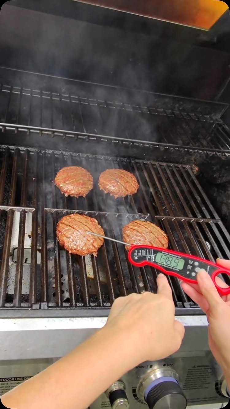 Red digital meat thermometer checking the temperature of beef patties on a grill