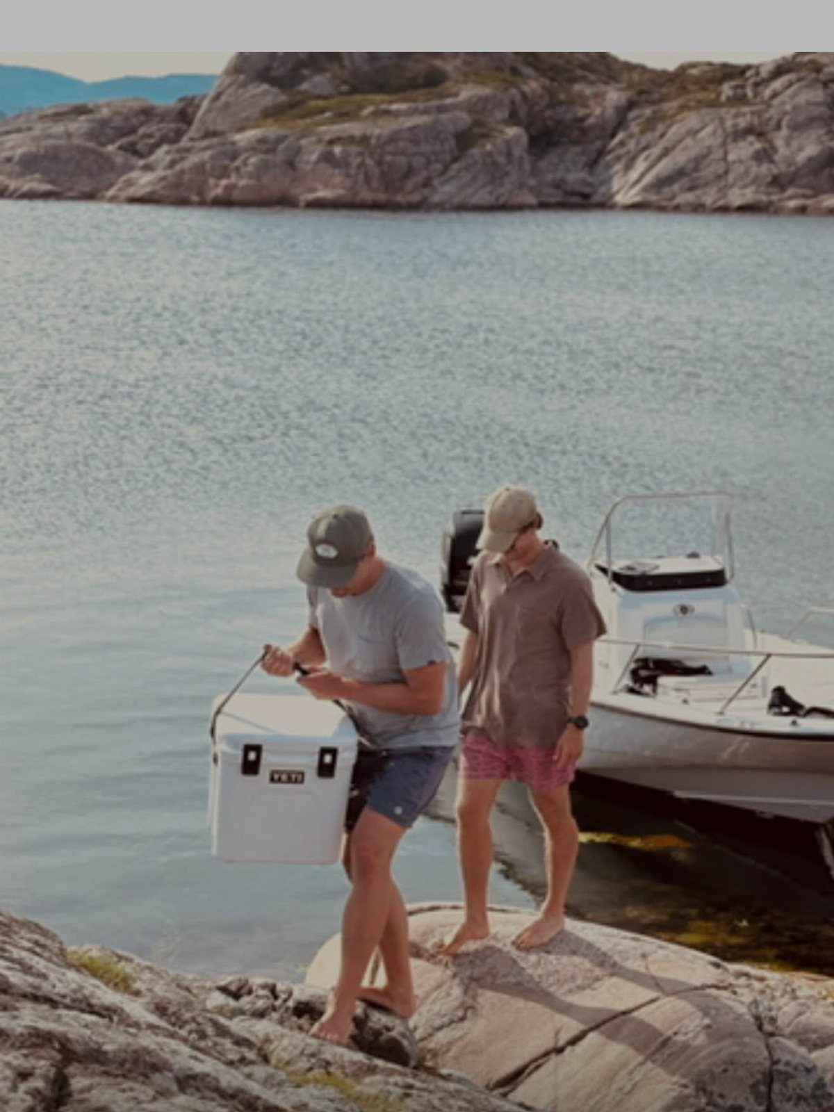 Two men on a rocky shore with a white YETI hard cooler and boat.