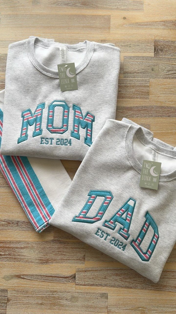 Two grey sweatshirts with MOM and DAD printed in striped letters on wood.