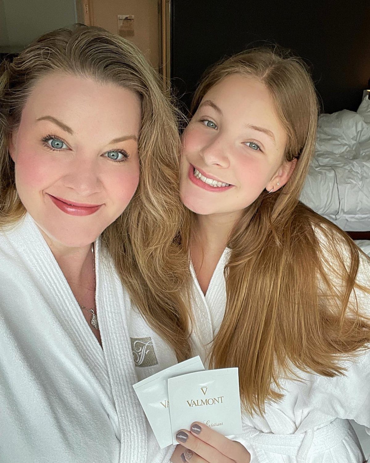 Two women in white bathrobes smiling and holding skincare packets for a spa day.