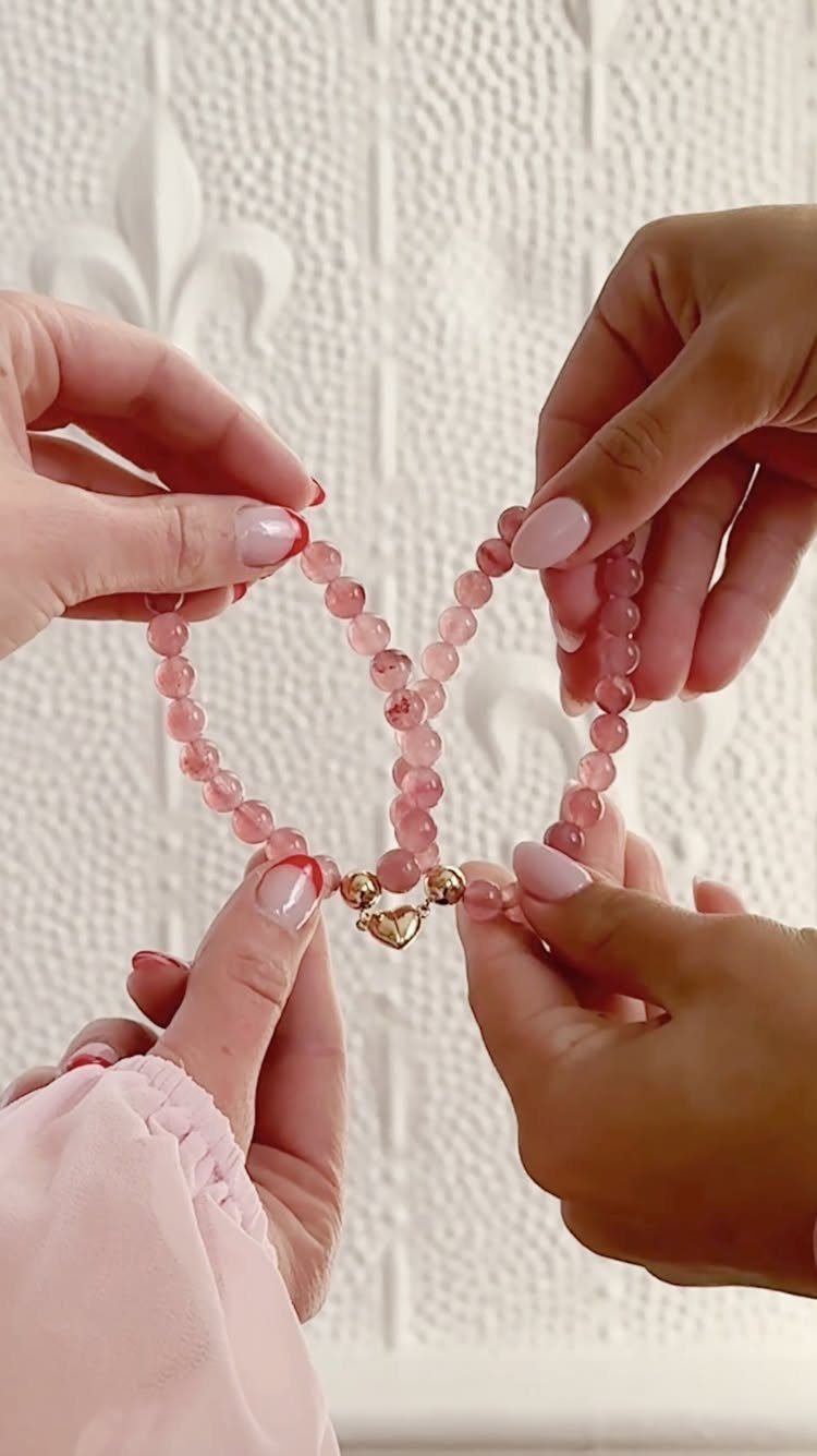 Two pink beaded bracelets joined by a gold heart charm held by hands.