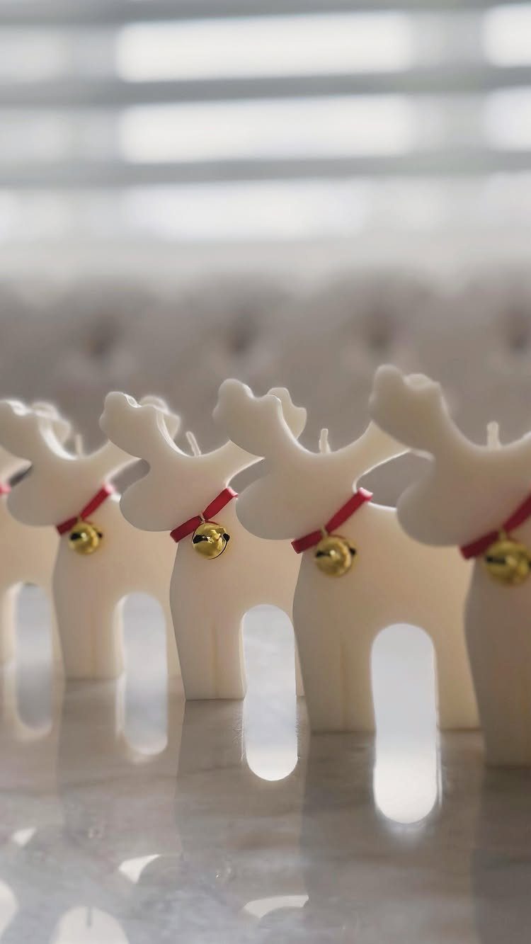 White reindeer candles with red collars and gold bells on a reflective surface.