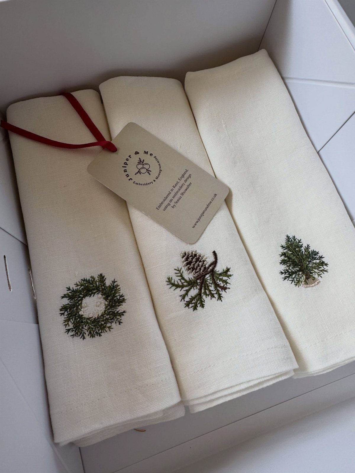 Cream fabric napkins with embroidered green Christmas wreaths and pinecones in a white box