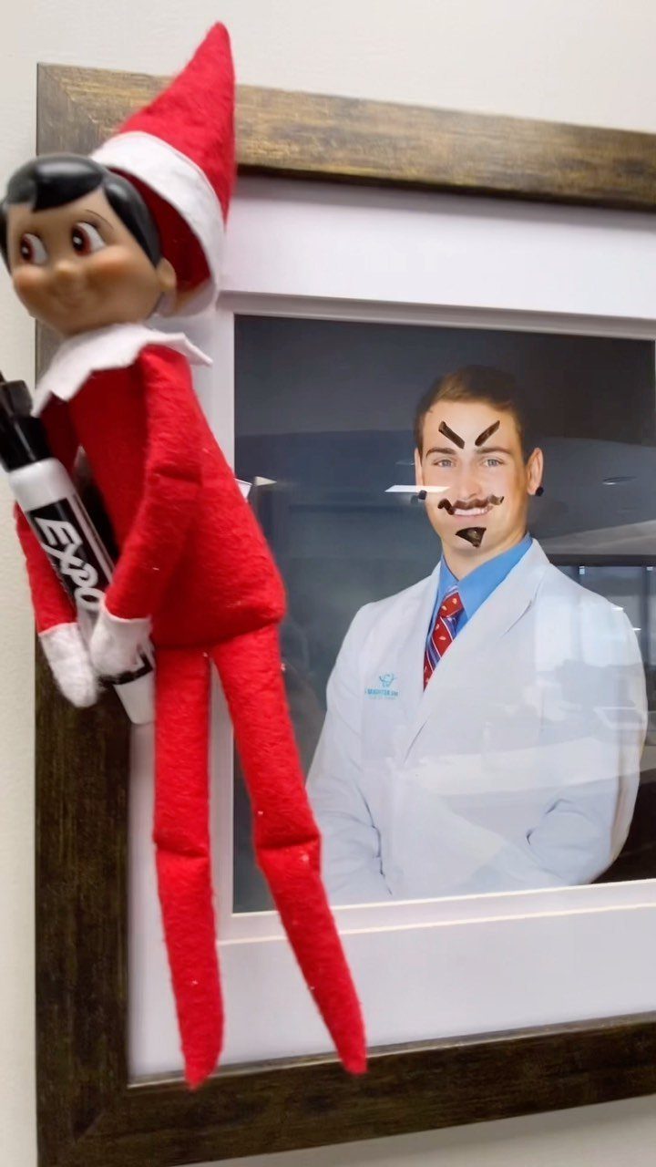 Elf on the Shelf doll with a marker sitting on a framed portrait.
