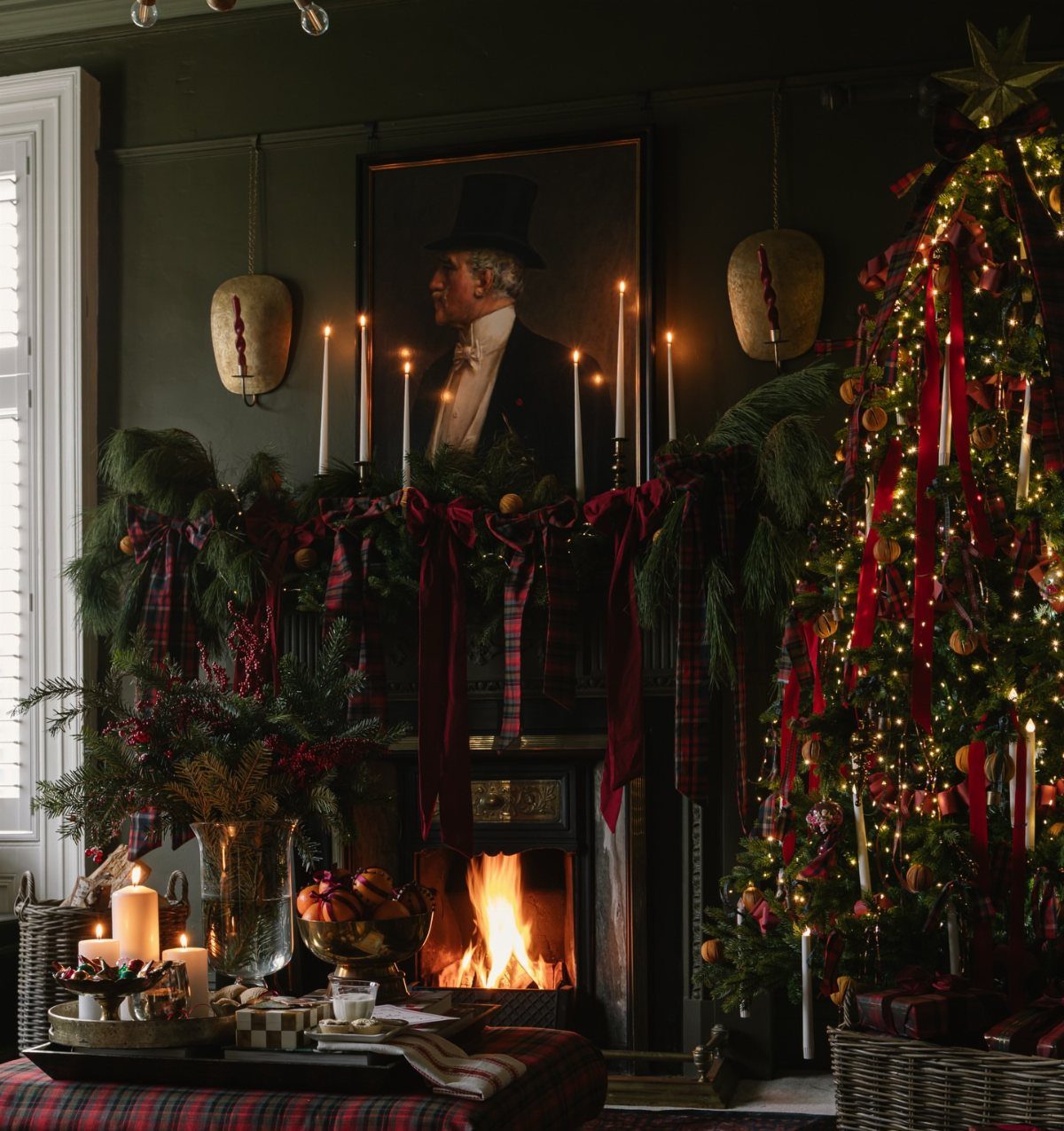 Fireplace mantel decorated with evergreen garlands, red plaid ribbons, and glowing candles.