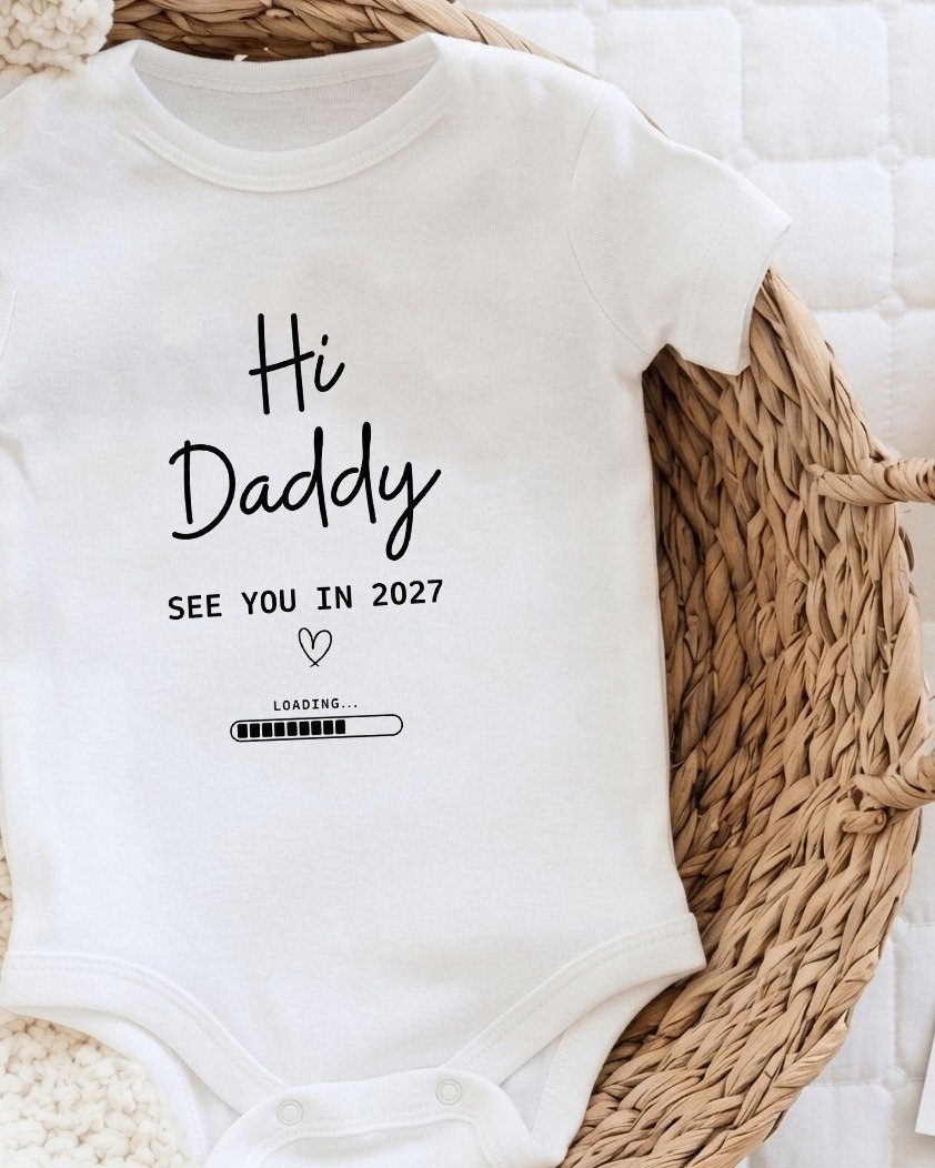 White baby bodysuit with Hi Daddy text resting on a wicker basket