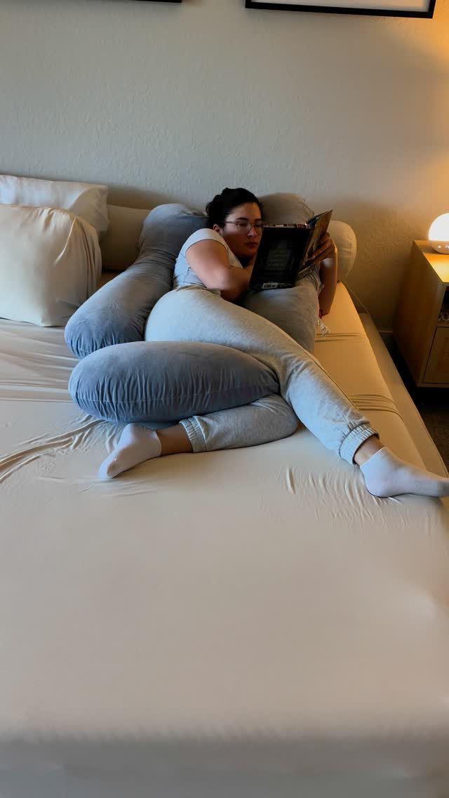 Pregnant woman reading a book while resting on a grey U-shaped pregnancy pillow