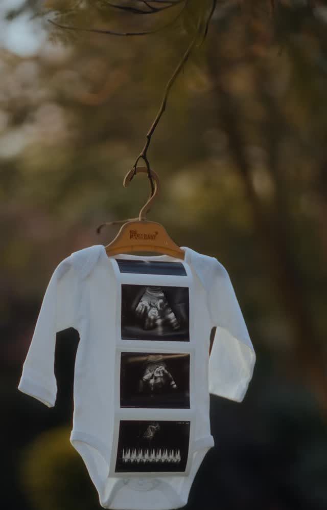 White baby bodysuit with ultrasound photos hanging from a wooden hanger outdoors