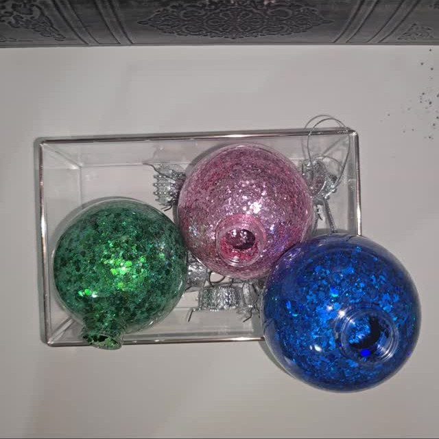 Three glittery Christmas ornaments in green, pink, and blue colors near a clear box.