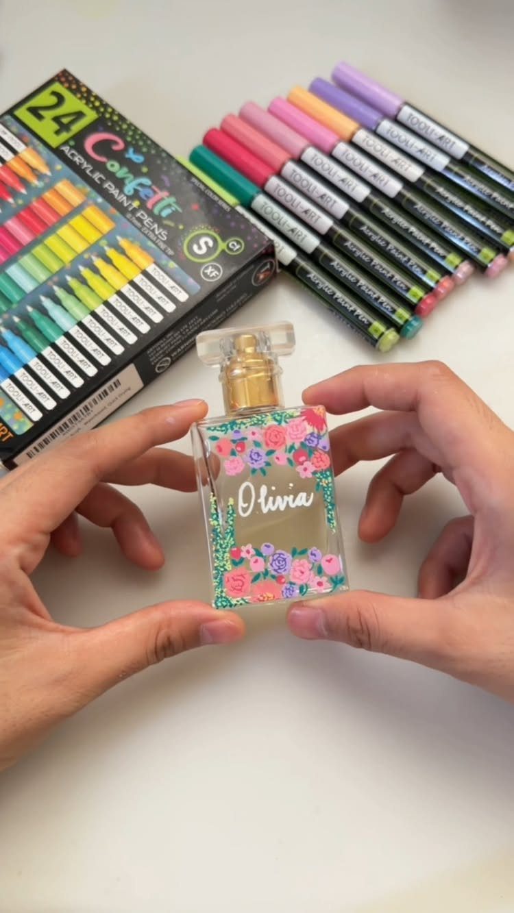 Hands holding a perfume bottle painted with flowers and a name using paint pens.