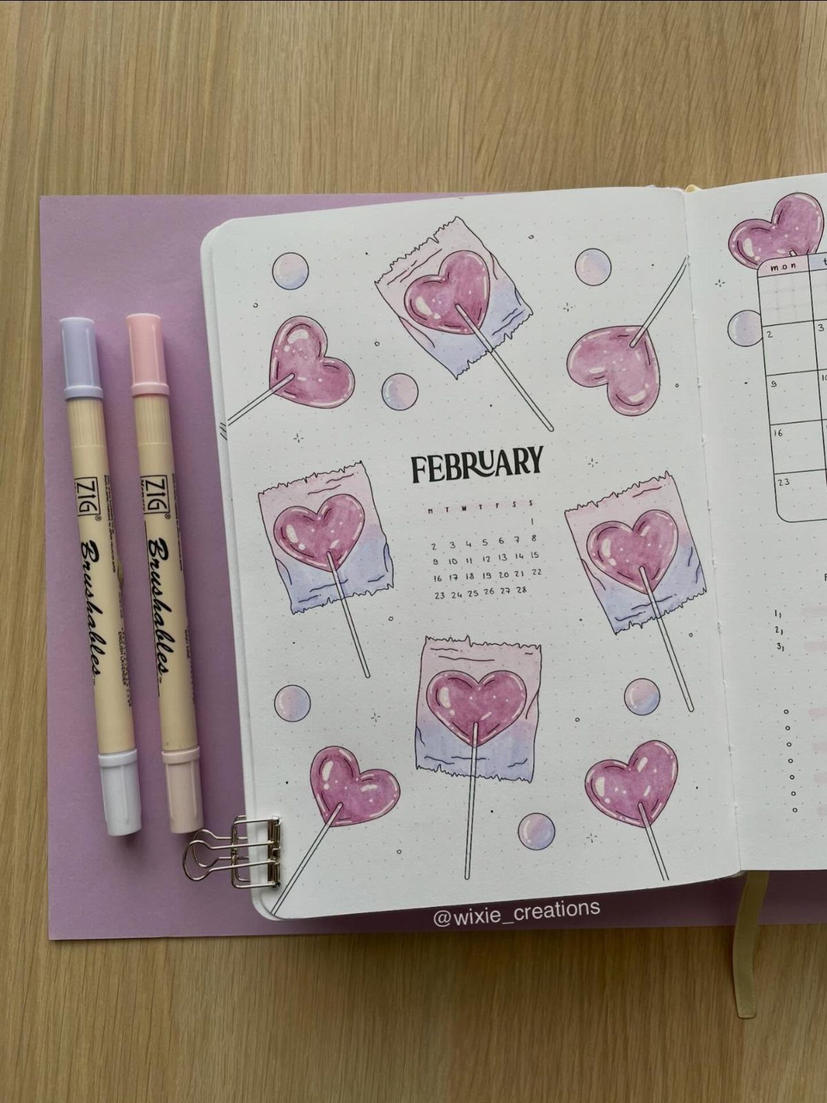 Open bullet journal with February calendar and pink heart lollipop drawings and pens.