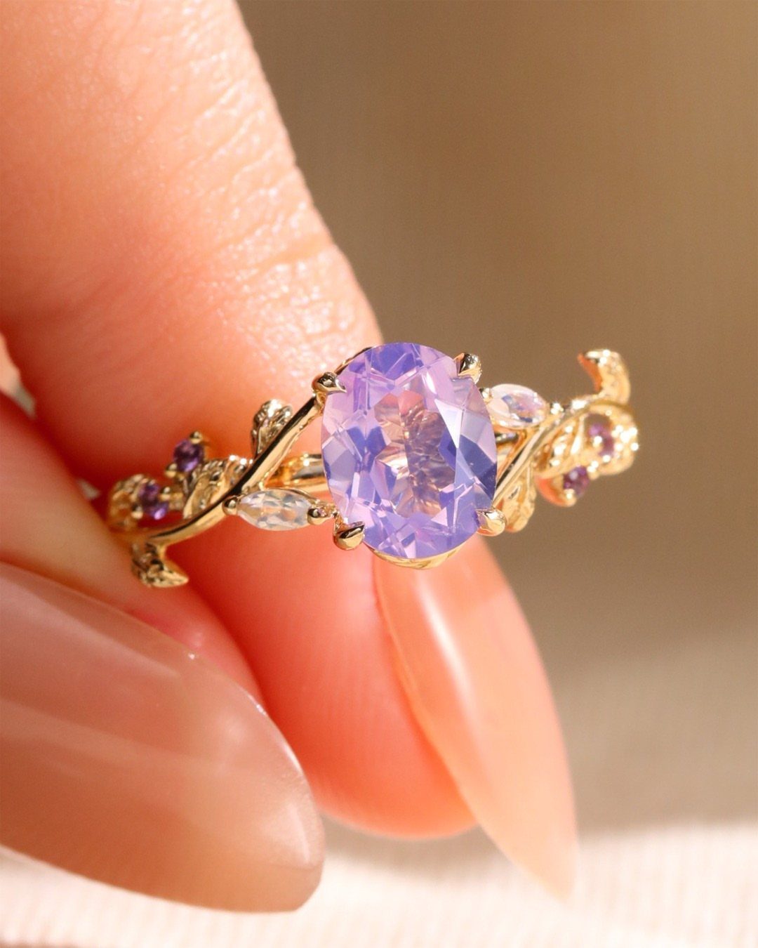 Gold ring with oval purple gemstone and leaf design held by fingers.