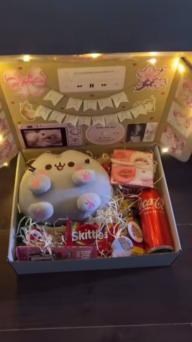 Gift box with Pusheen plush, candy, and soda decorated with fairy lights.