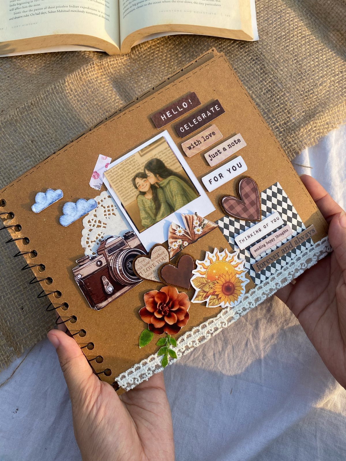 Hands holding a brown spiral scrapbook decorated with photos, flowers, and vintage stickers.